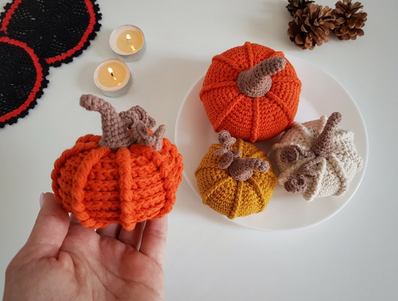 May include: Four crocheted pumpkins in orange, yellow, and cream with brown stems. The pumpkins are on a white plate.