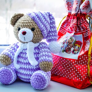 May include: A crocheted teddy bear wearing a purple and white hat and matching outfit. The bear is sitting next to a red gift bag with a white snowflake pattern.