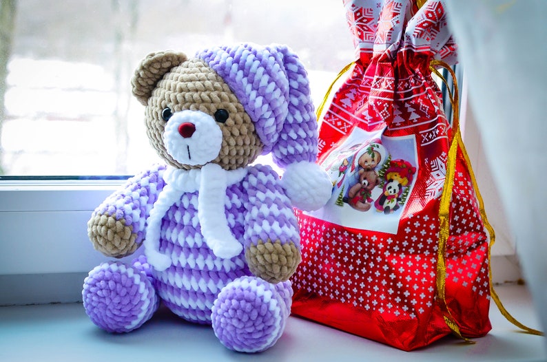 May include: A crocheted teddy bear wearing a purple and white hat and matching outfit. The bear is sitting next to a red gift bag with a white snowflake pattern.