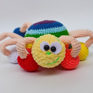 Pu&ograve; includere: Un giocattolo ragno all'uncinetto colorato con un viso giallo peloso, occhi neri e un sorriso verde. Il ragno ha otto zampe, ciascuna terminante con una palla all'uncinetto di diversi colori: viola, bianco, rosso, giallo e verde.