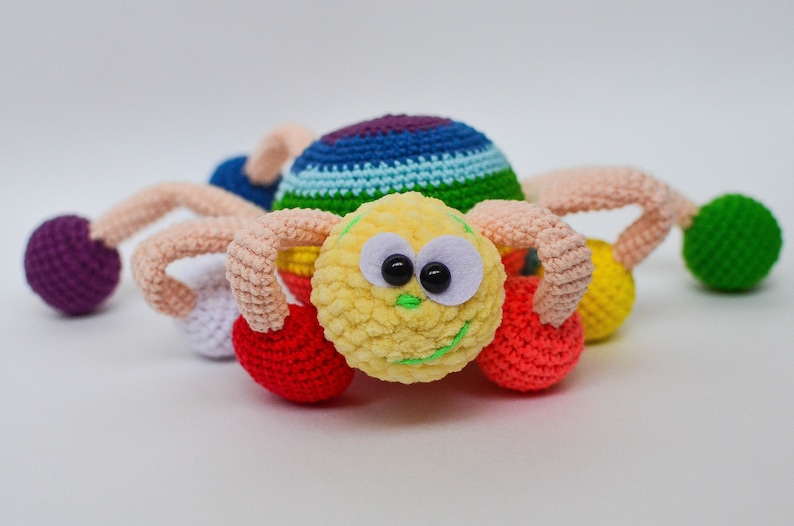 Pu&ograve; includere: Un giocattolo ragno all'uncinetto colorato con un viso giallo peloso, occhi neri e un sorriso verde. Il ragno ha otto zampe, ciascuna terminante con una palla all'uncinetto di diversi colori: viola, bianco, rosso, giallo e verde.