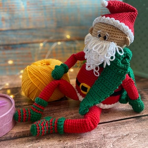 May include: A crocheted Santa Claus doll with a red hat, white beard, and green scarf. The doll is sitting on a wooden surface with a ball of yellow yarn.