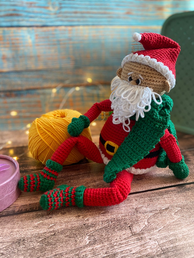 May include: A crocheted Santa Claus doll with a red hat, white beard, and green scarf. The doll is sitting on a wooden surface with a ball of yellow yarn.
