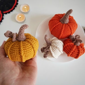 May include: Four crocheted pumpkins in different colors and sizes. The pumpkins are on a white plate. The pumpkins are orange, yellow, and white.