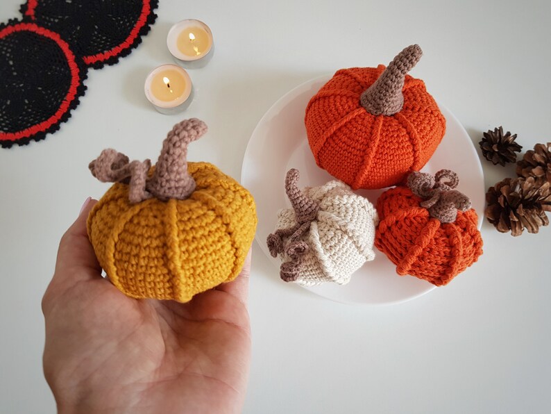 May include: Four crocheted pumpkins in different colors and sizes. The pumpkins are on a white plate. The pumpkins are orange, yellow, and white.