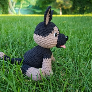 May include: A crocheted stuffed dog toy in black and brown sitting on green grass. The dog has a black nose and a pink tongue sticking out.