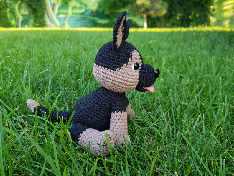 May include: A crocheted stuffed dog toy in black and brown sitting on green grass. The dog has a black nose and a pink tongue sticking out.