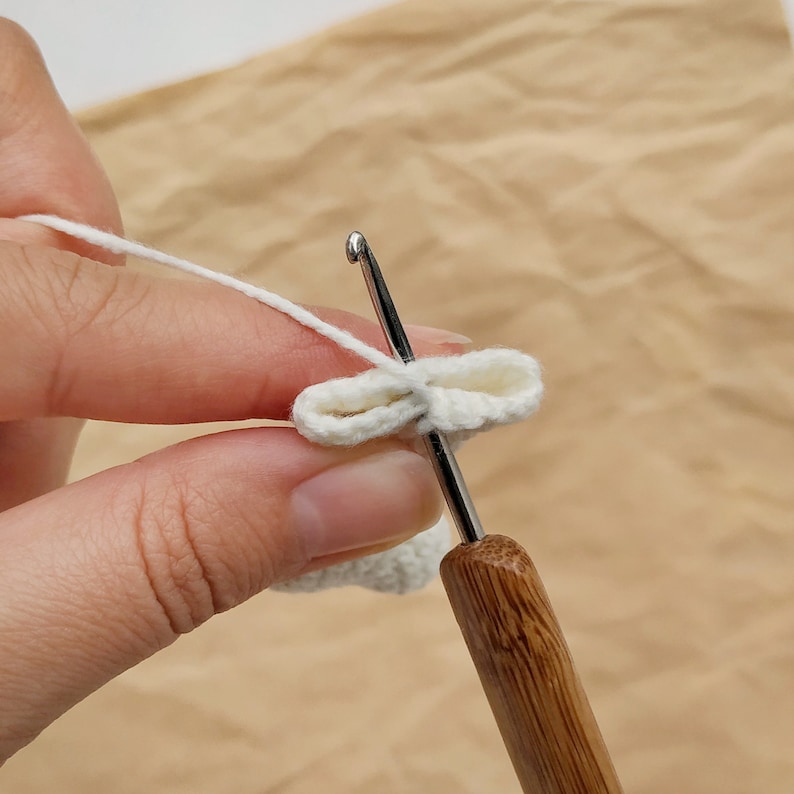 May include: A close-up of a white crocheted piece being worked on with a crochet hook. The hook is made of wood and has a metal hook.