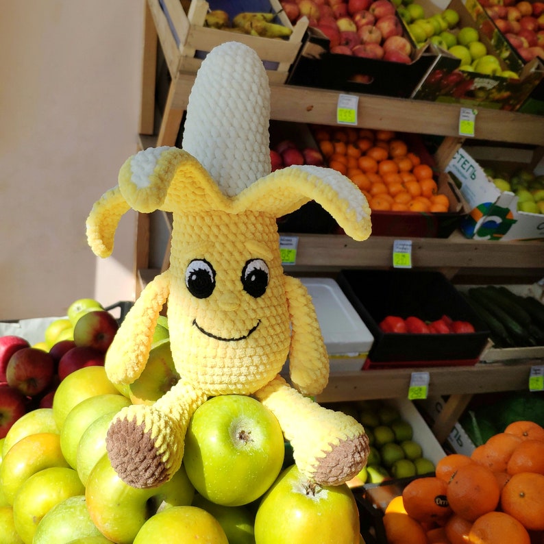 May include: A crocheted banana plush toy with a smiling face. The toy is sitting on a pile of green apples in front of a wooden shelf with fruit and vegetables.