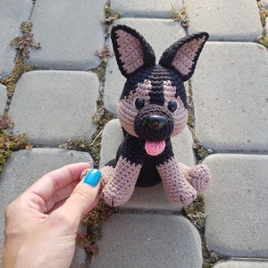 May include: A crocheted plush toy dog with black and brown fur, pink tongue, and black eyes. The dog is sitting on a gray stone surface.