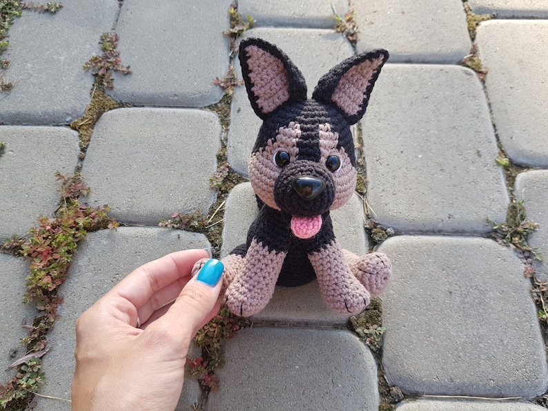 May include: A crocheted plush toy dog with black and brown fur, pink tongue, and black eyes. The dog is sitting on a gray stone surface.