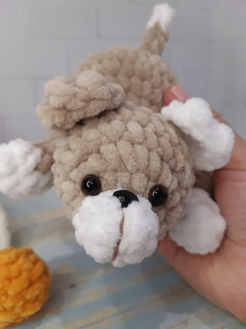May include: A crocheted plush toy dog with brown and white fur, black eyes, and a black nose. The dog is lying on its side with its head tilted up.