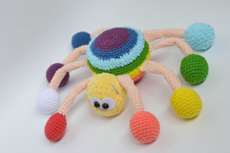 Pu&ograve; includere: Un giocattolo ragno all'uncinetto colorato con un corpo a strisce arcobaleno e otto gambe, ciascuna terminante con una palla di colore diverso. Il ragno ha occhietti buffi e una testa gialla pelosa.