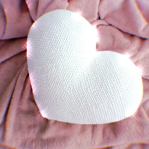 May include: A white crocheted heart-shaped pillow on a pink fuzzy surface.
