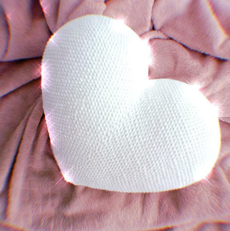 May include: A white crocheted heart-shaped pillow on a pink fuzzy surface.