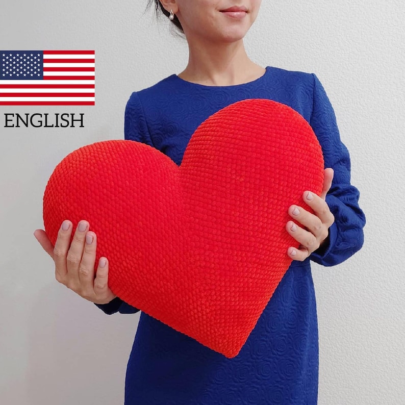 May include: A large, red, heart-shaped pillow held by a person. The pillow is made of a soft, textured fabric.