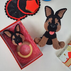 K&ouml;nnte beinhalten: Zwei geh&auml;kelte Spielzeug-Deutscher Sch&auml;ferhunde. Einer ist ein gr&ouml;&szlig;eres Pl&uuml;schtier und der andere ist ein kleineres Rasselspielzeug in einer roten Schachtel mit rosa Seidenpapier.