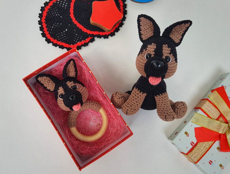 K&ouml;nnte beinhalten: Zwei geh&auml;kelte Spielzeug-Deutscher Sch&auml;ferhunde. Einer ist ein gr&ouml;&szlig;eres Pl&uuml;schtier und der andere ist ein kleineres Rasselspielzeug in einer roten Schachtel mit rosa Seidenpapier.