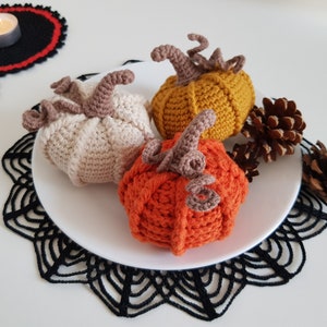 May include: Three crocheted pumpkins in orange, beige, and yellow on a white plate with black lace doilies and pine cones.