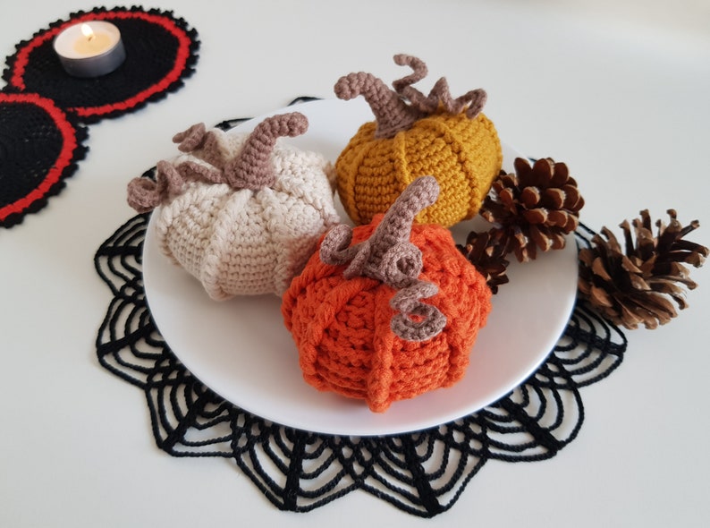 May include: Three crocheted pumpkins in orange, beige, and yellow on a white plate with black lace doilies and pine cones.