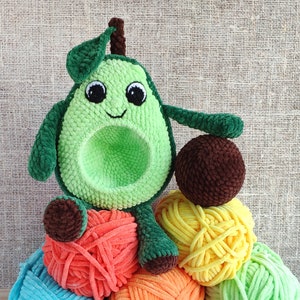 Puede incluir: Un juguete de peluche de aguacate tejido a crochet con una cara sonriente, sentado sobre una pila de ovillos de lana de colores. El aguacate es verde con un hueso marrón y tiene un tallo tejido a crochet marrón.