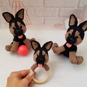 K&ouml;nnte beinhalten: Drei geh&auml;kelte Spielzeug-Deutscher Sch&auml;ferhunde, eines mit einem Holz-Bei&szlig;ring. Die Hunde sind schwarz und braun mit rosa Zungen. Ein Hund sitzt auf einem roten Ball.
