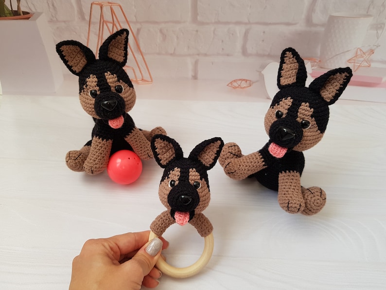 K&ouml;nnte beinhalten: Drei geh&auml;kelte Spielzeug-Deutscher Sch&auml;ferhunde, eines mit einem Holz-Bei&szlig;ring. Die Hunde sind schwarz und braun mit rosa Zungen. Ein Hund sitzt auf einem roten Ball.