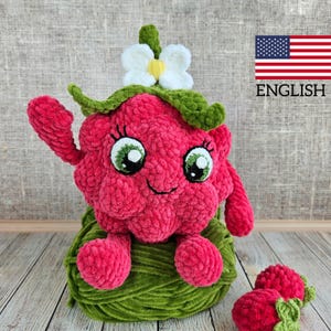 Peut inclure: Un jouet en peluche en forme de fraise crocheté avec une fleur blanche sur le dessus, assis sur une pelote de laine verte. La fraise a un visage souriant avec des yeux verts et un corps rose. Il y a deux plus petites fraises crochetées au premier plan.