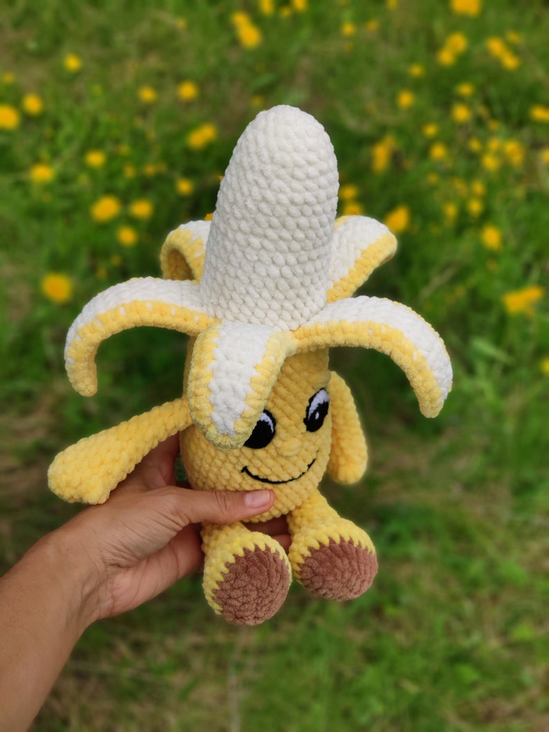May include: A crocheted banana plush toy with a smiling face and brown feet. The banana is yellow with a white crocheted top.