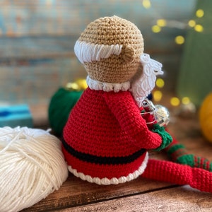May include: Crocheted Santa Claus doll with a red suit, white trim, and a white beard. The doll is holding a silver ornament in its hand.