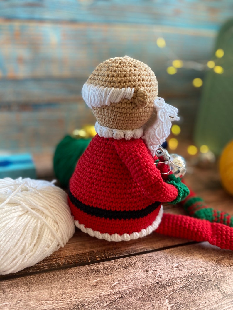 May include: Crocheted Santa Claus doll with a red suit, white trim, and a white beard. The doll is holding a silver ornament in its hand.