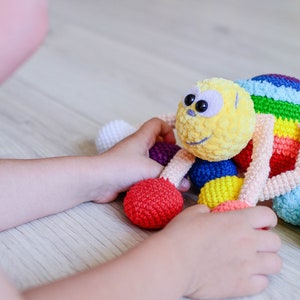 Pu&ograve; includere: Un giocattolo ragno all'uncinetto colorato con una testa gialla e pelosa e grandi occhi neri. Il ragno ha gambe arcobaleno e &egrave; circondato da palline all'uncinetto colorate.