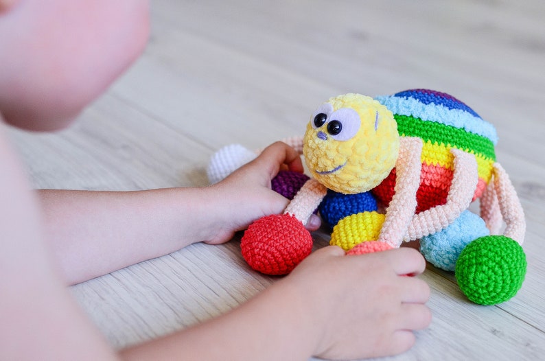 Pu&ograve; includere: Un giocattolo ragno all'uncinetto colorato con una testa gialla e pelosa e grandi occhi neri. Il ragno ha gambe arcobaleno e &egrave; circondato da palline all'uncinetto colorate.