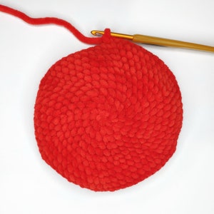 May include: A red crocheted round cushion with a textured surface. The cushion is being worked on with a crochet hook and yarn.