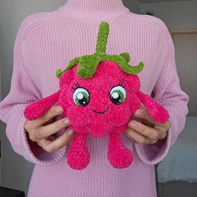 May include: A pink crocheted raspberry plush toy with green leaves and a smiling face. The toy has large, round eyes and a small, stitched mouth.
