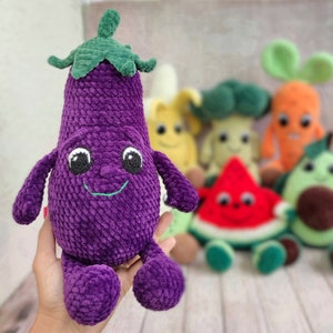 Puede incluir: Un juguete de peluche de berenjena de crochet morado con un tallo verde y una cara sonriente. La berenjena está rodeada de otros juguetes de peluche de verduras de crochet, incluyendo una zanahoria, una sandía, un brócoli, un plátano y un aguacate.