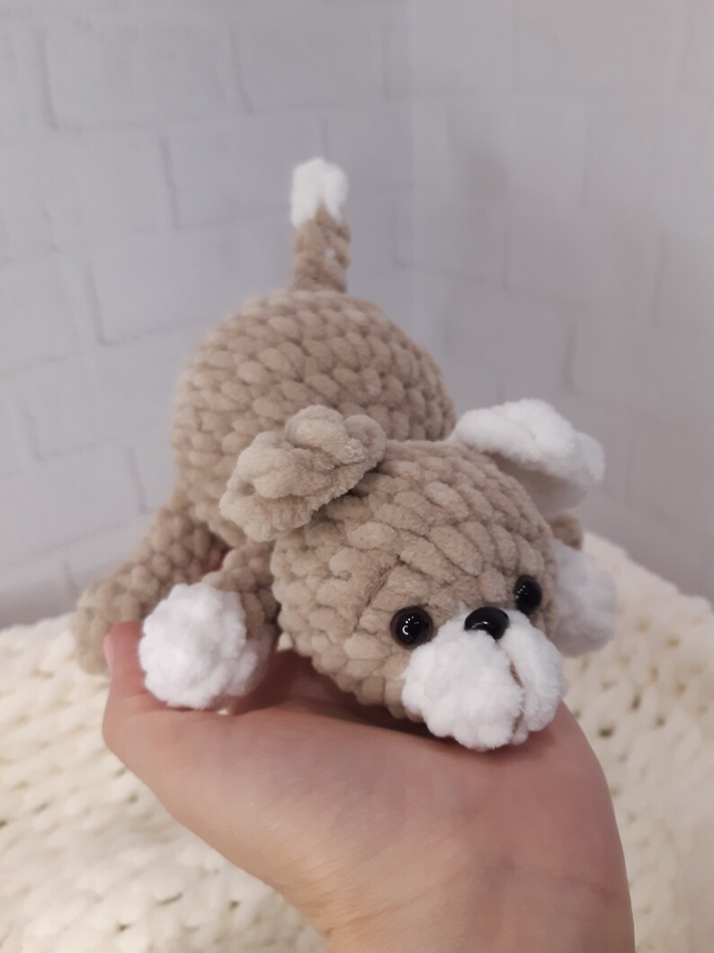 May include: A crocheted plush toy dog with brown fur and white accents. The dog has black eyes and a white nose.