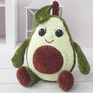 K&ouml;nnte beinhalten: Ein Pl&uuml;sch-Avocado-Spielzeug mit einem braunen Kern, einem gr&uuml;nen K&ouml;rper und einem freundlichen Gesicht. Die Avocado hat ein kleines gr&uuml;nes Blatt oben drauf.
