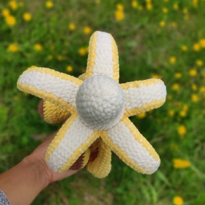 May include: A crocheted plush toy shaped like a banana with a white and yellow striped pattern. The toy is held in a hand against a blurred background of yellow flowers.