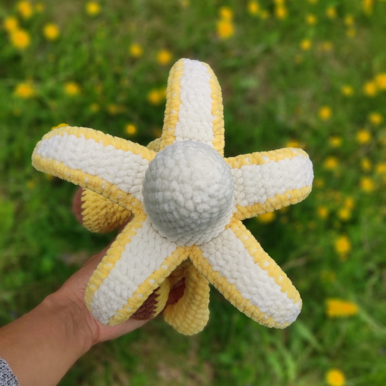 May include: A crocheted plush toy shaped like a banana with a white and yellow striped pattern. The toy is held in a hand against a blurred background of yellow flowers.