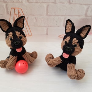 May include: Two crocheted German Shepherd dog plushies, one brown and black with a pink tongue sticking out, sitting on a white surface with a red ball in front of it. The other plush is brown and black with a pink tongue sticking out.