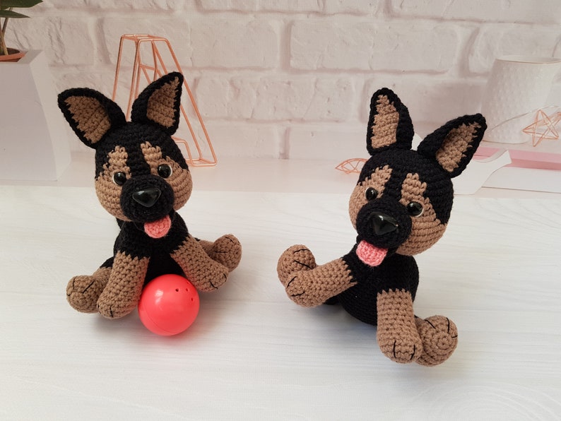 May include: Two crocheted German Shepherd dog plushies, one brown and black with a pink tongue sticking out, sitting on a white surface with a red ball in front of it. The other plush is brown and black with a pink tongue sticking out.