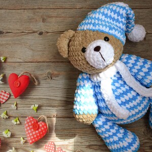 May include: A crocheted teddy bear wearing a blue and white sleep cap and pajamas. The bear is lying on a wooden surface with red and white hearts scattered around it.