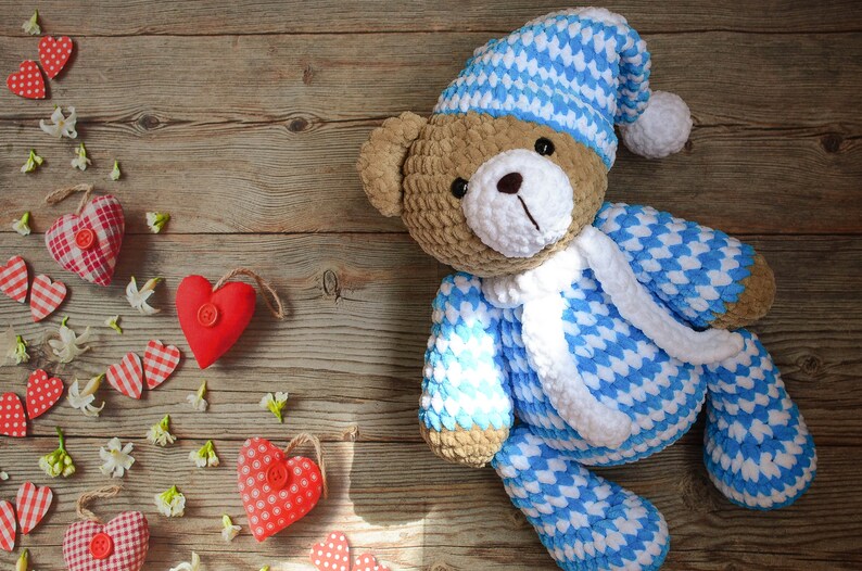 May include: A crocheted teddy bear wearing a blue and white sleep cap and pajamas. The bear is lying on a wooden surface with red and white hearts scattered around it.