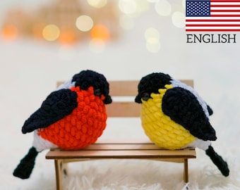 May include: Two crocheted birds, one red and one yellow, are sitting on a wooden bench. The birds have black and white accents.
