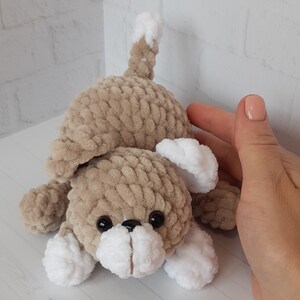 May include: A crocheted plush toy dog with brown fur, white paws, and black eyes. The dog is lying on its belly with its head tilted up.