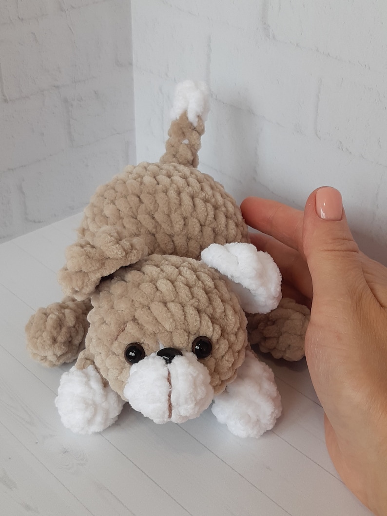 May include: A crocheted plush toy dog with brown fur, white paws, and black eyes. The dog is lying on its belly with its head tilted up.