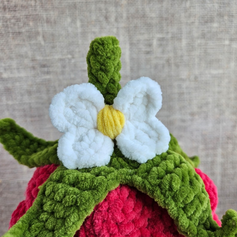 May include: A crocheted strawberry with green leaves and a white flower with a yellow center.