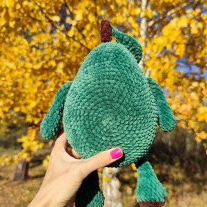 K&ouml;nnte beinhalten: Ein gr&uuml;nes, geh&auml;keltes Avocado-Pl&uuml;schtier mit braunen Filzdetails. Das Spielzeug wird in einer Hand mit pinkfarbenem Nagellack vor einem Hintergrund aus gelben Herbstbl&auml;ttern gehalten.