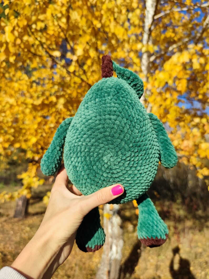 K&ouml;nnte beinhalten: Ein gr&uuml;nes, geh&auml;keltes Avocado-Pl&uuml;schtier mit braunen Filzdetails. Das Spielzeug wird in einer Hand mit pinkfarbenem Nagellack vor einem Hintergrund aus gelben Herbstbl&auml;ttern gehalten.
